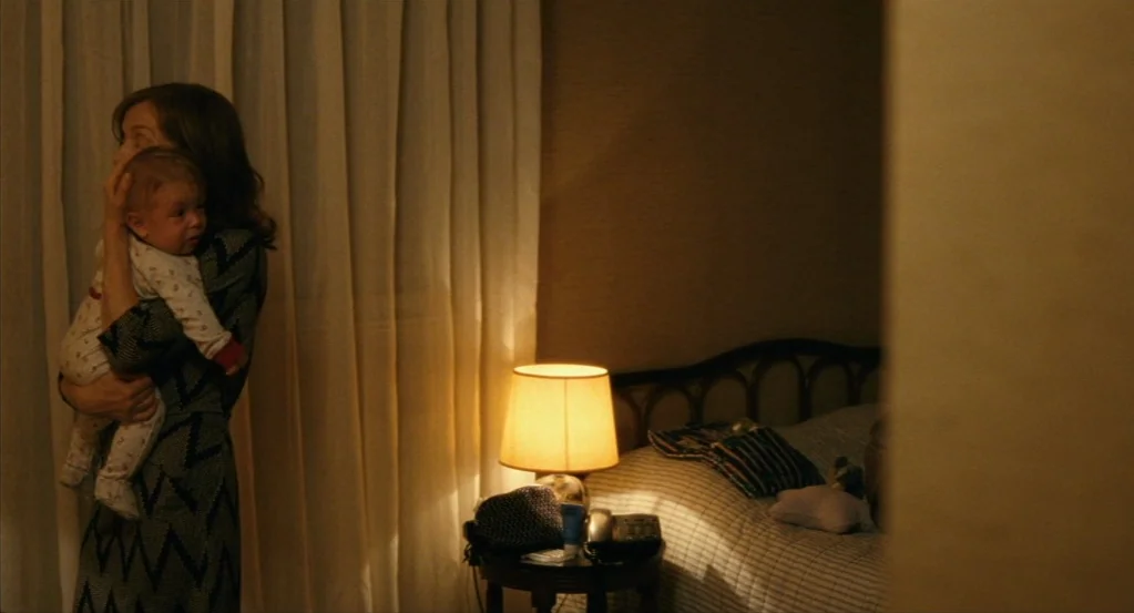 Things to Come (2016) movie still: medium — A dark hotel room features a rumpled bed and a bedside lamp with curtains. A woman in a patterned dress…