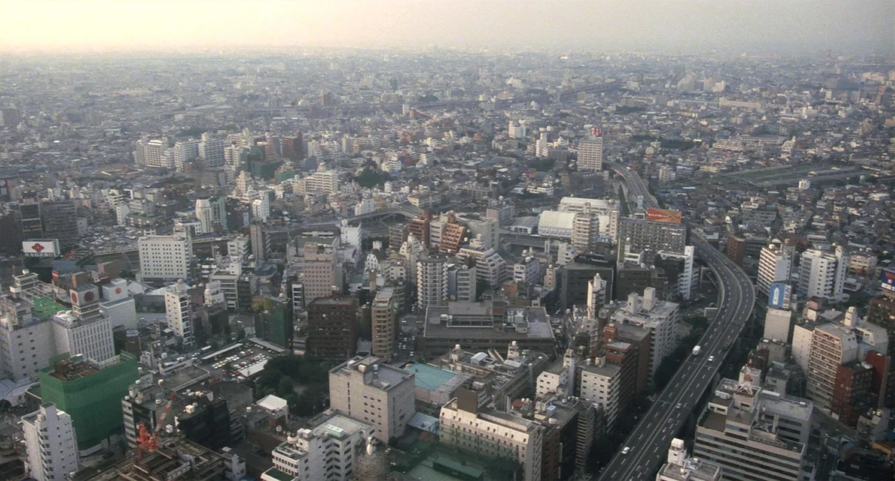 Tampopo (1985) movie still: establishing — A dense urban expanse of Tokyo with mid-rise buildings and a curving highway. The city extends to a hazy…