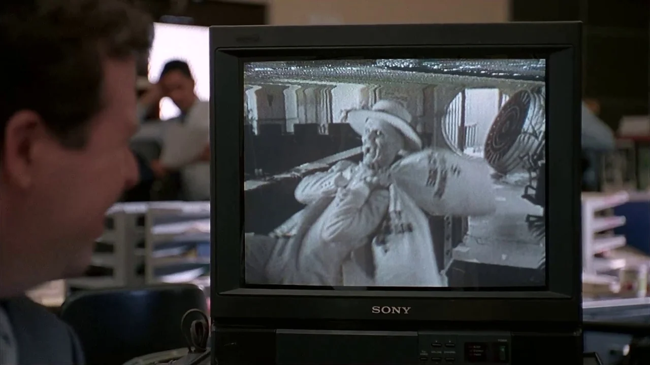 The Mask (1994) movie still: medium — A man in a professional setting sits at a desk viewing a monitor. The screen displays a grainy,…
