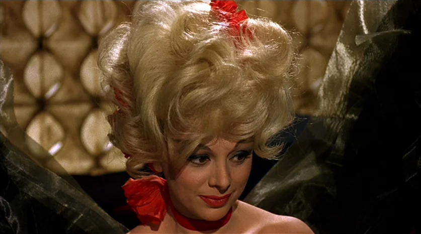 Juliet of the Spirits (1965) movie still: closeup — A woman with a tall blonde bouffant is centered in a dimly lit space. She gazes downward while…