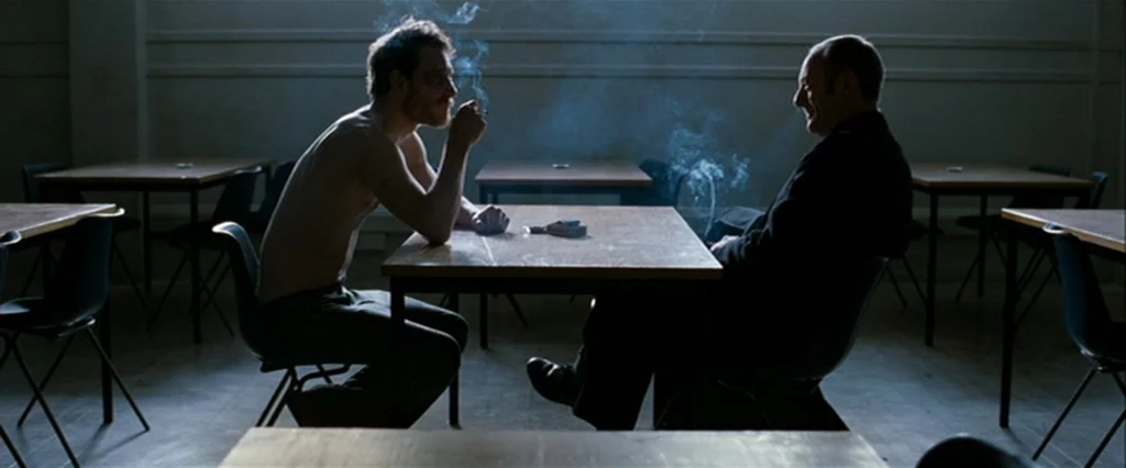 Hunger (2008) movie still: wide — A sparse cafeteria with wooden tables and black chairs arranged in rows. Two men sit at a central table, smoking…