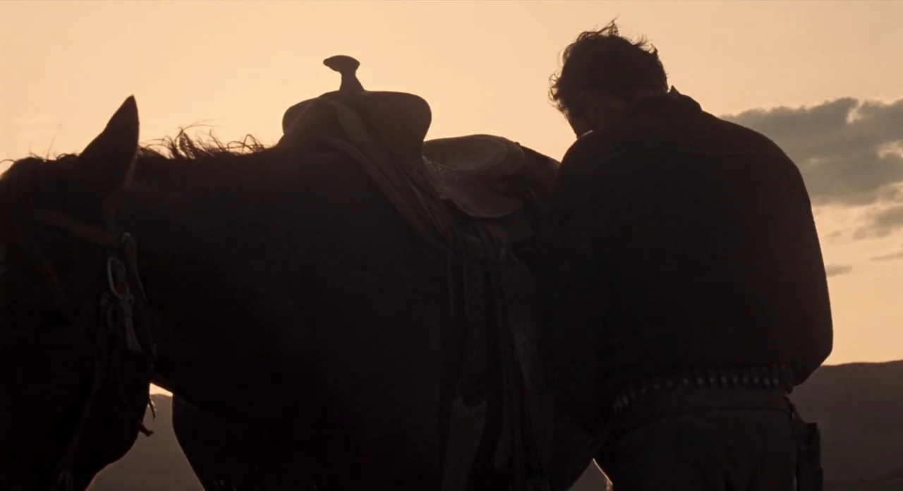 The Shooting (1966) movie still: medium — An open plain during a sunset with a horse and man. The man stands by the horse tightening a saddle cinch.