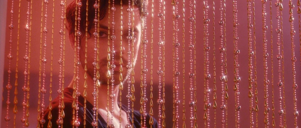 Tamara Drewe (2010) movie still: closeup — A man stands behind a dense screen of hanging metallic gold and silver beads. Intense pink light washes…