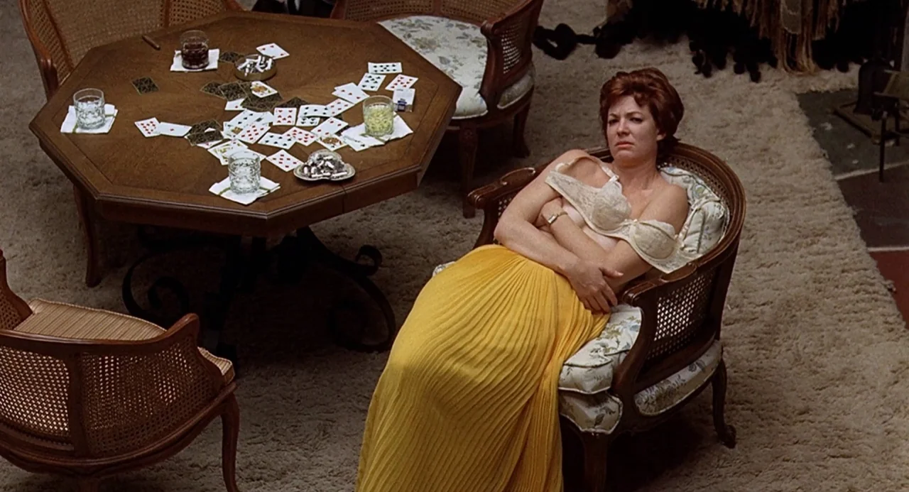 Taking Off (1971) movie still: medium — A living room with an octagonal wooden game table cluttered with playing cards. A woman sits in a patterned…