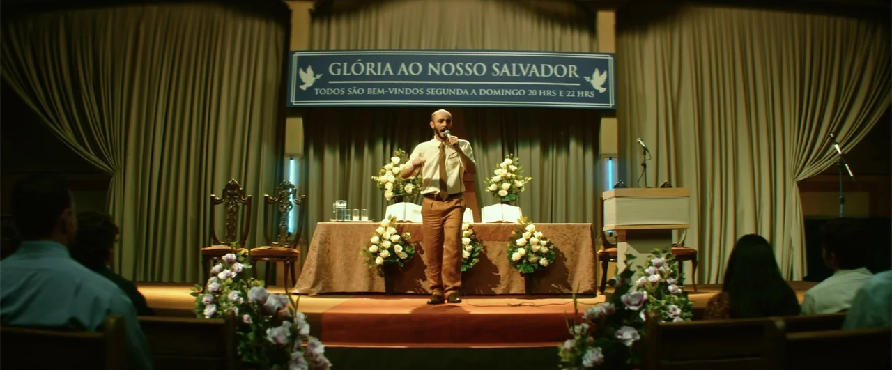 Bingo: The King of the Mornings (2017) movie still: fullbody — A small church stage features a banner in Portuguese draped over heavy tan curtains.…