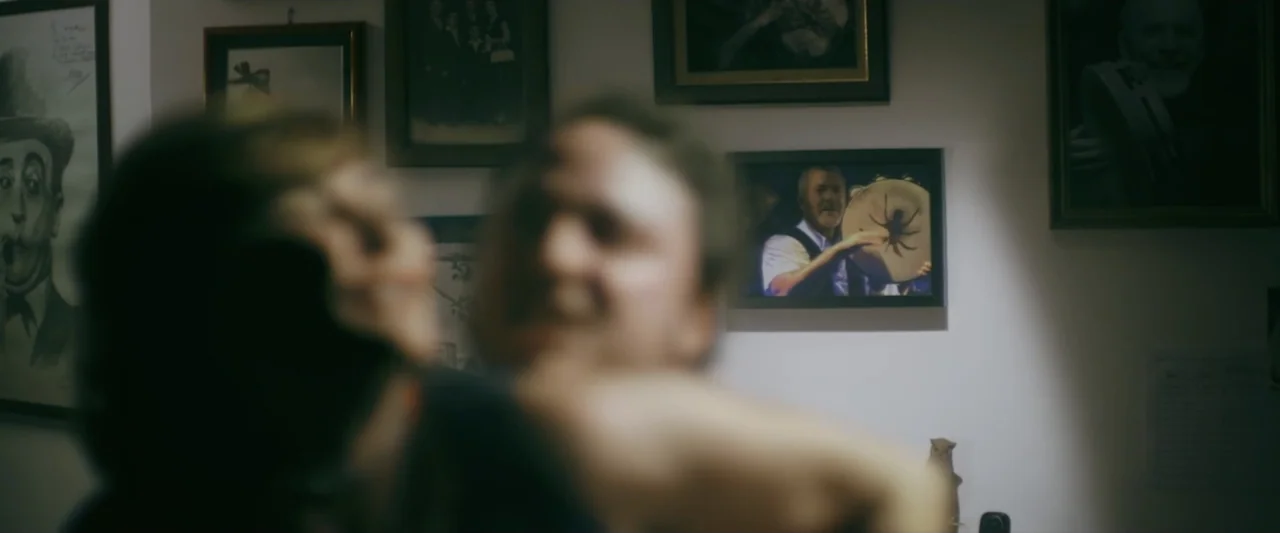 Taranta On the Road (2017) movie still: medium — A wall is covered with multiple framed photographs and portraits. Two people are in the foreground,…