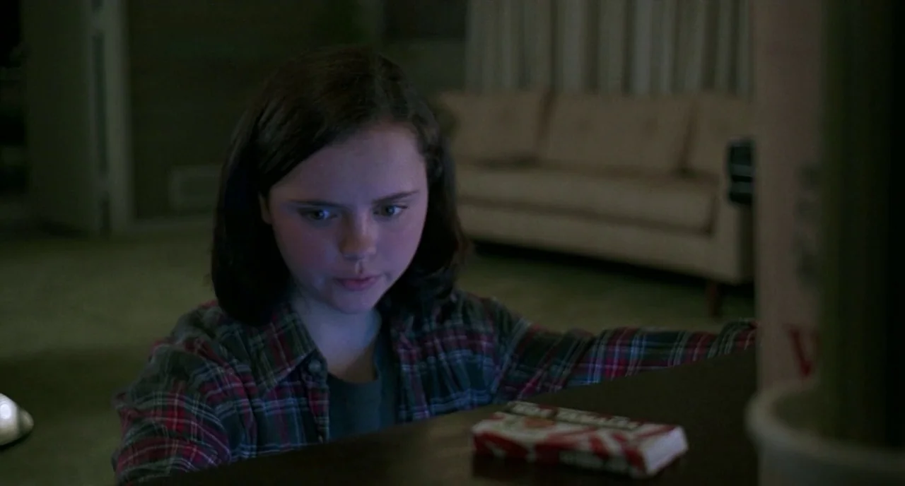 The Ice Storm (1997) movie still: medium — A dimly lit room contains a sofa in the background and a television setup. A young girl stares at a candy…