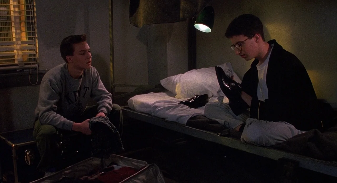 Child’s Play 3 (1991) movie still: medium — A spartan dorm room with a metal frame bed and a suitcase on the floor. Two young men are present in the…