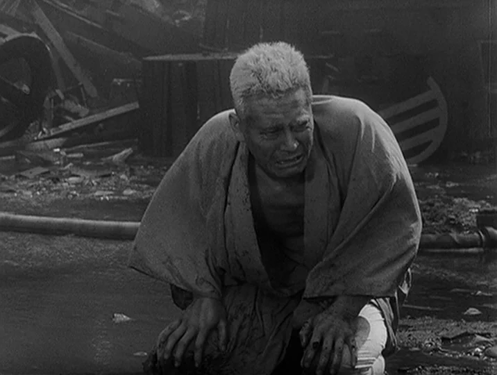 I Live In Fear (1955) movie still: medium — An outdoor area covered in wreckage and twisted debris from a destroyed building. A middle-aged man…