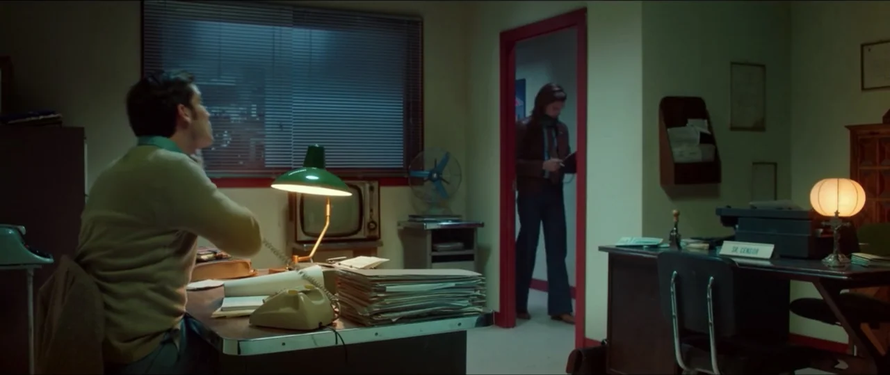 My Heart Goes Boom! (2020) movie still: medium — A cluttered administrative office features green walls, two desks, and a window with blinds. One…