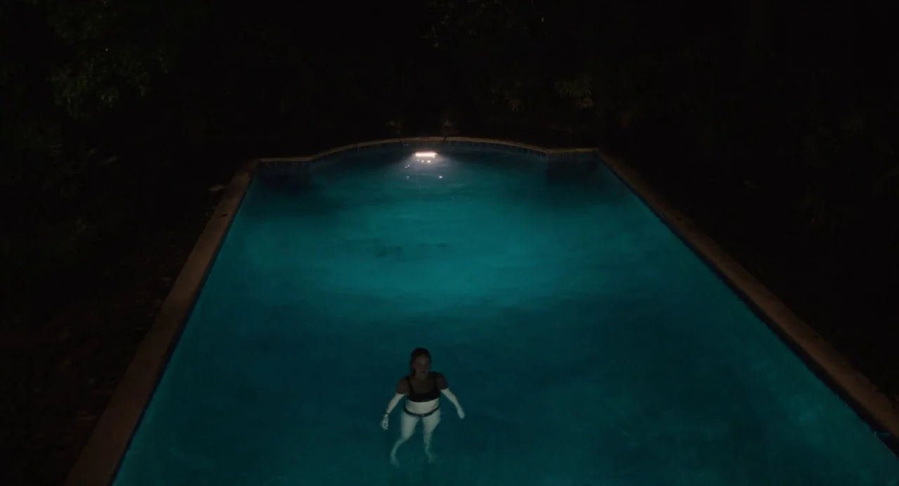 Causeway movie still: wide — A rectangular swimming pool surrounded by dark trees is lit by an underwater lamp. A lone woman swims in the center of…