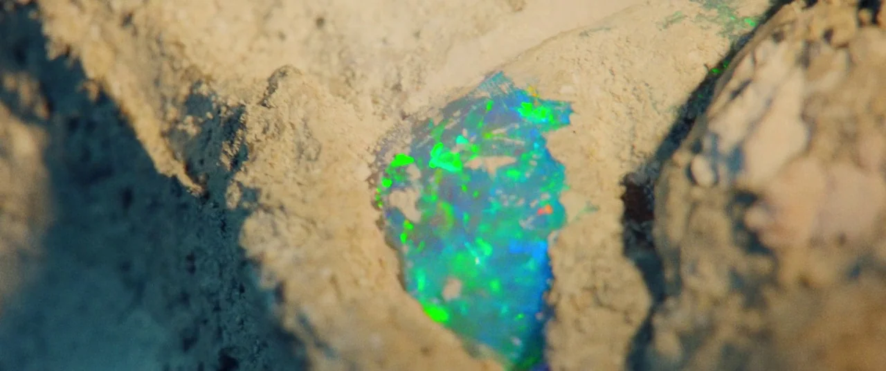 Uncut Gems (2019) movie still: closeup — A raw, unrefined opal is embedded within a jagged, sandy-colored rock formation. The gem displays bright…