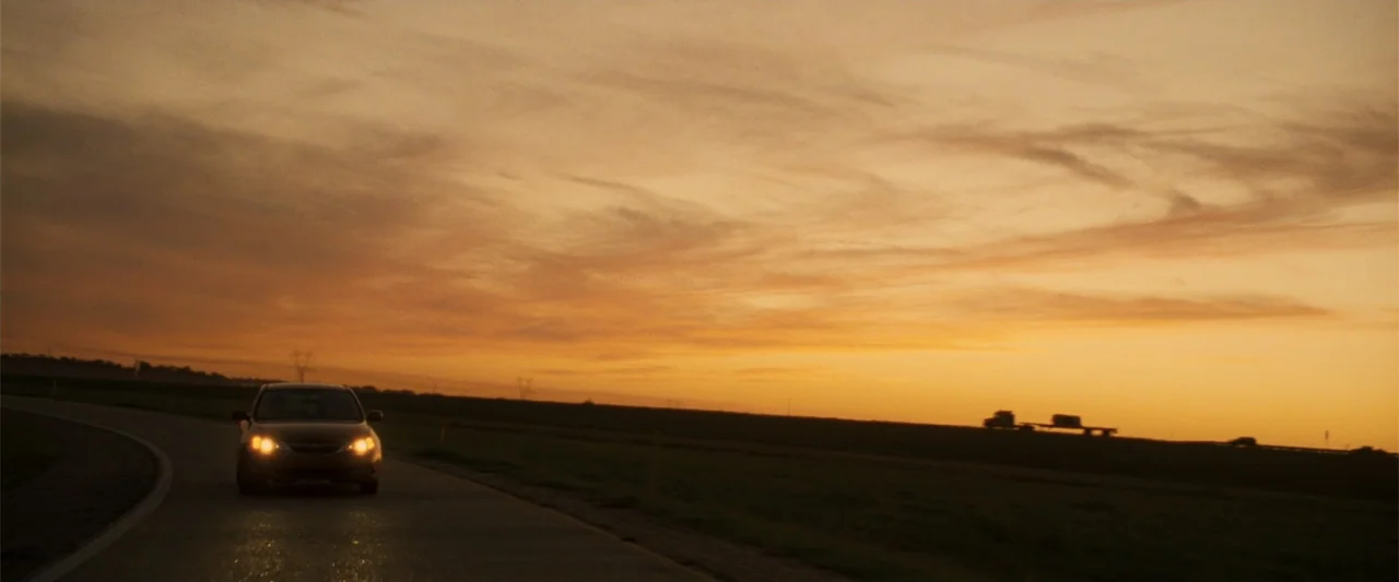 Due Date (2010) movie still: wide — A flat highway stretches across a rural landscape during a clear golden hour sunset. A single sedan drives…