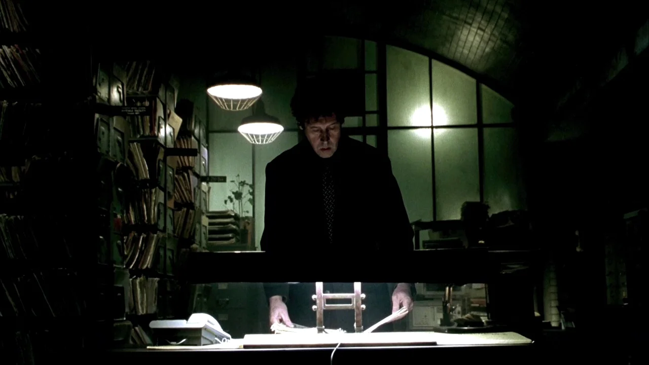In Dreams (1999) movie still: medium — A dark, cramped room is lined with tall, packed metal shelving units filled with files. A man in a dark suit…