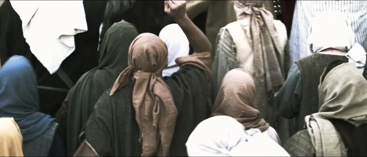 The Penitent Thief (2021) movie still: medium — A dense group of people in simple draped garments fills an outdoor public space. A central figure…