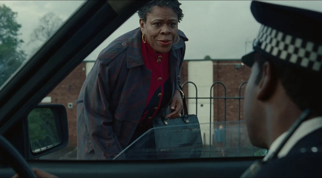 Red, White & Blue (2020) movie still: medium — An older woman stands outside a car, speaking through an open window to an officer. A brick wall with…