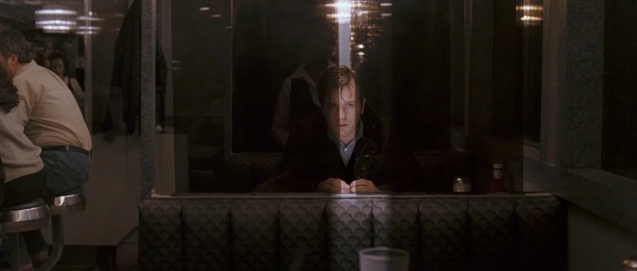 Stay (2005) movie still: medium — A man sits alone in a diner booth behind a large glass pane at night. The reflective surface bisects his face with…