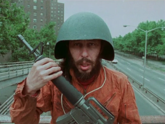 WR: Mysteries of the Organism (1971) movie still: medium — A man in a jumpsuit stands on a pedestrian bridge above a highway. He holds a black rifle…