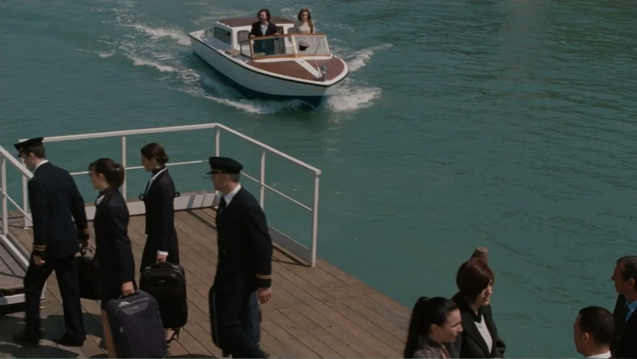 The Tourist (2010) movie still: wide — A wooden pier extends over turquoise water with a motorboat moving toward the landing. Airline crew members…