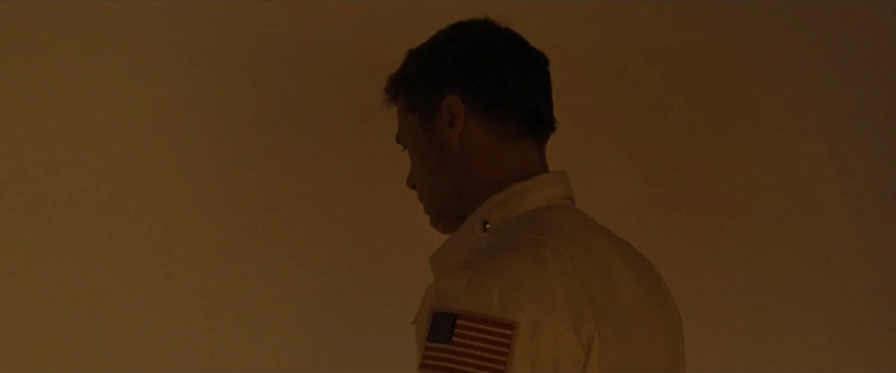 Ad Astra (2019) movie still: medium — A dimly lit interior space with plain walls surrounds a man from behind. A man with short hair wearing a white…