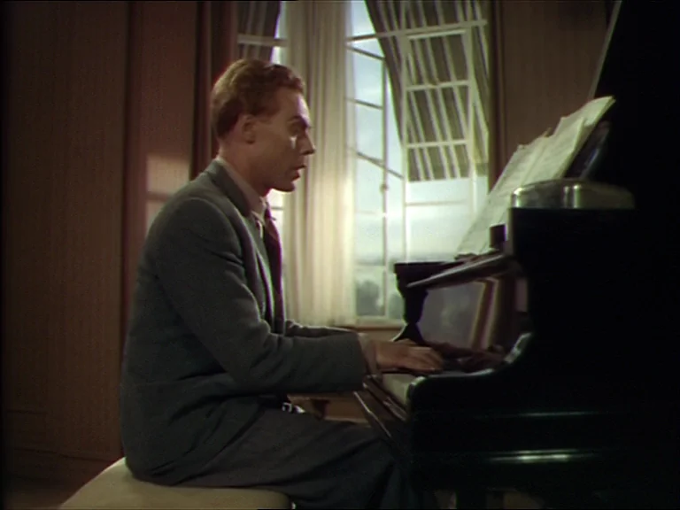 The Red Shoes (1948) movie still: medium — A dimly lit room features a large window with sheer white curtains. A man sits at a black piano playing…