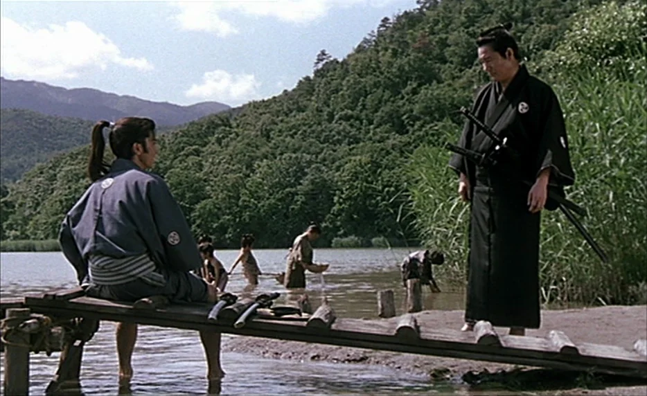 Gohatto (1999) movie still: wide — A wooden dock extends into a calm lake surrounded by lush green forested hills. A man sits on the edge while…
