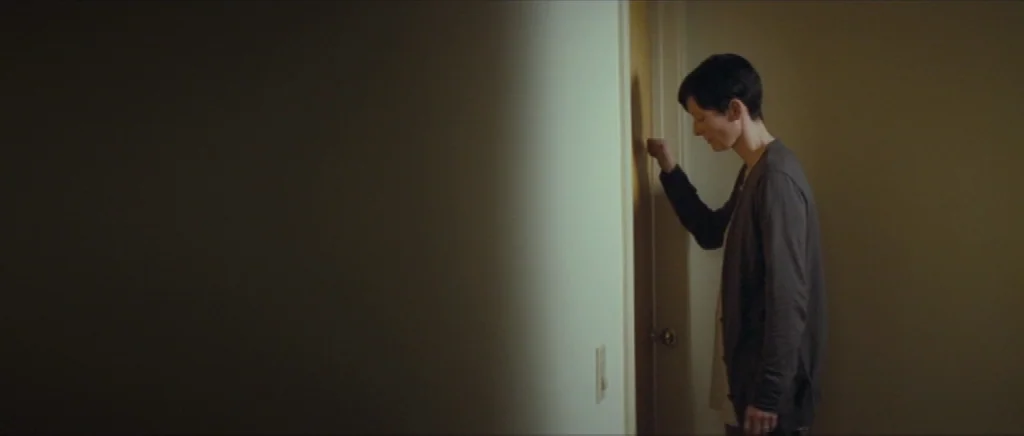 We Need To Talk About Kevin (2011) movie still: medium — A dimly lit indoor hallway features beige walls and a single closed wooden door. A woman…
