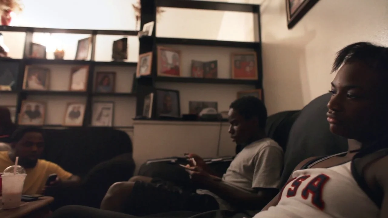 T-Rex (2015) movie still: medium — A dimly lit room features a large shelving unit filled with various framed photographs. Three young men sit on…