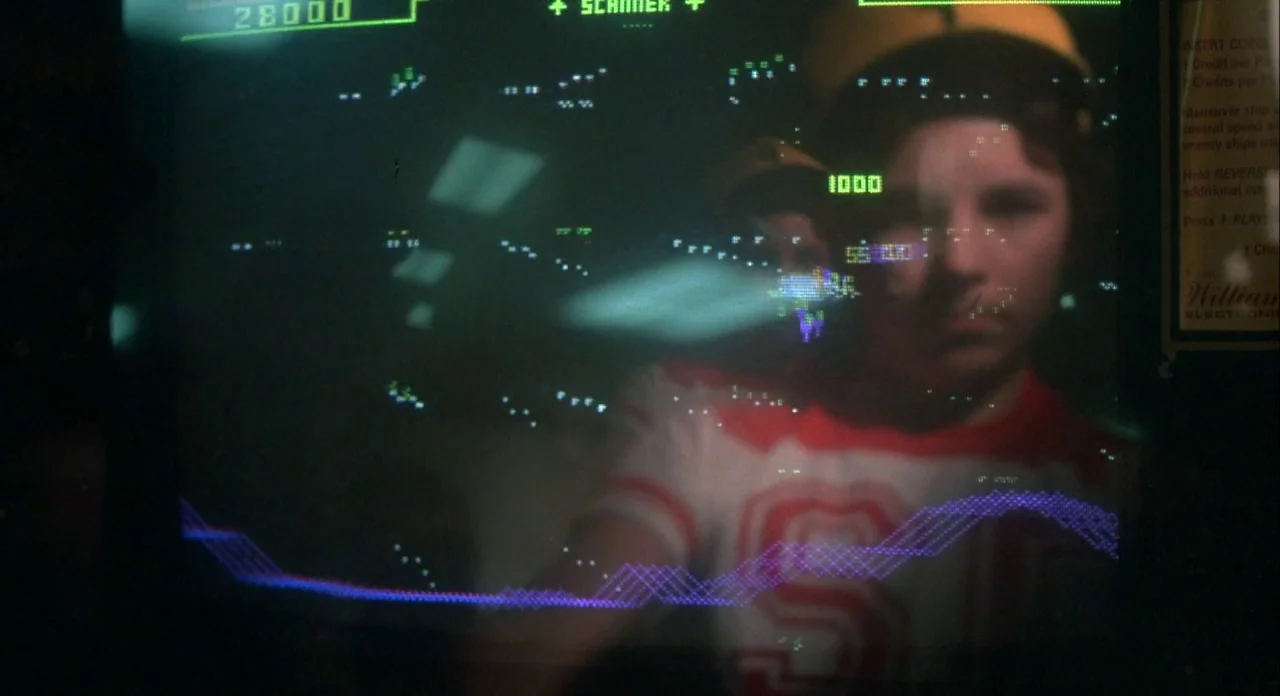 Koyaanasquatsi (1982) movie still: closeup — An arcade game monitor shows the reflection of a teenager wearing a yellow cap. The screen displays a…