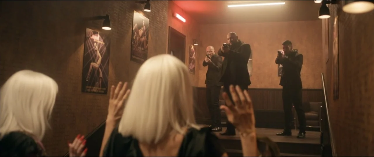 Risqué (2025) movie still: medium — A dark, wood-paneled room features promotional posters on the wall and a small stage. Three men point handguns…