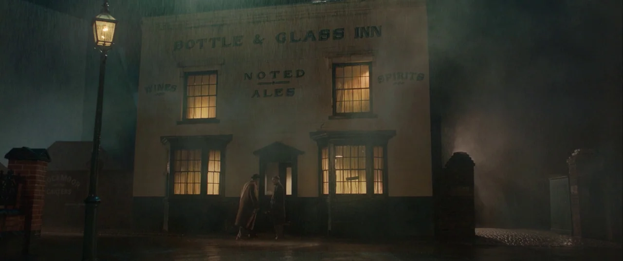 Domino (2005) movie still: wide — A two-story building labeled Bottle & Glass Inn stands on a street in heavy rain. Two men stand by the central…