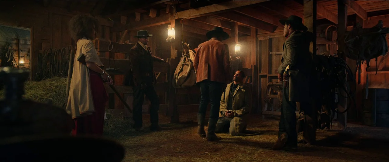 The Harder They Fall (2021) movie still: wide — The interior of a rustic wooden structure is dimly lit by hanging lanterns. A group of armed figures…