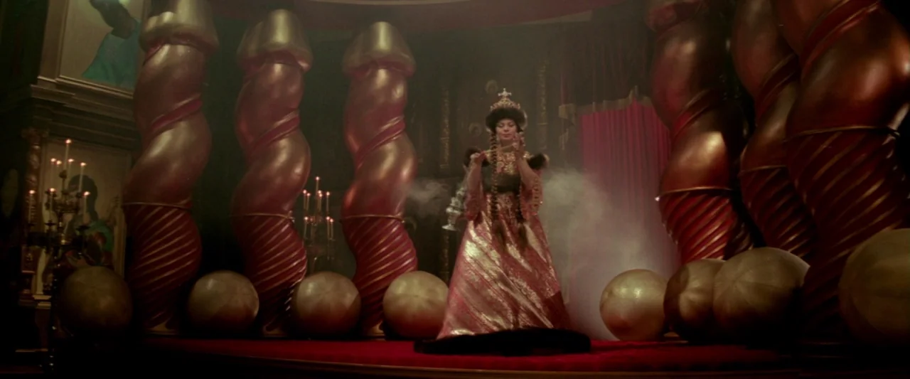 Lisztomania (1975) movie still: fullbody — A dark, opulent space features rows of large metallic spiral columns on a red dais. A lone woman dressed…