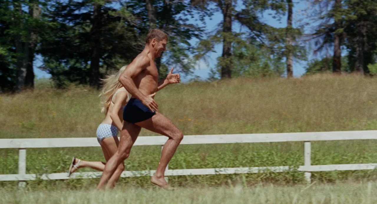 The Swimmer (1968) movie still: fullbody — A sunny grassy field with scattered trees and a white wooden rail fence. A shirtless man and a woman in a…