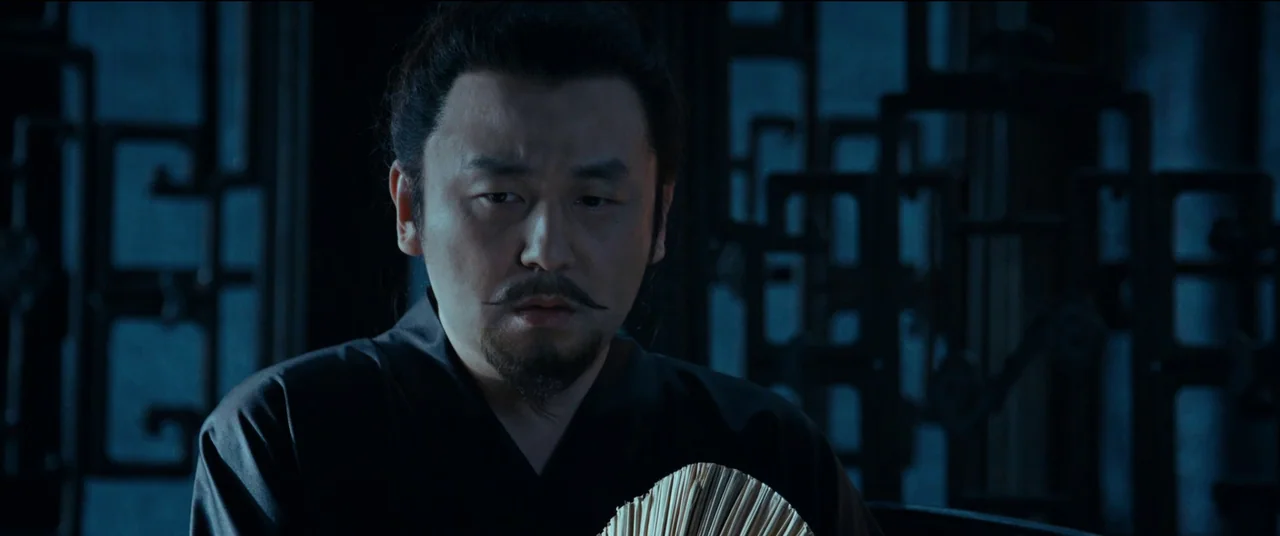 Full River Red (2023) movie still: closeup — A dark indoor space features decorative wooden latticework panels in the background. A man dressed in…