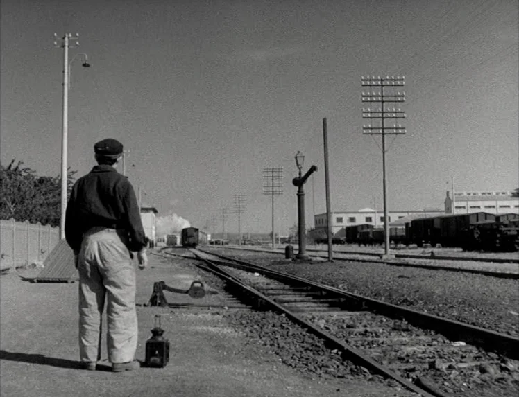 I Vitelloni (1953) movie still: fullbody — A desolate railway yard with metal tracks and telegraph poles stretching toward the horizon. A man in a…
