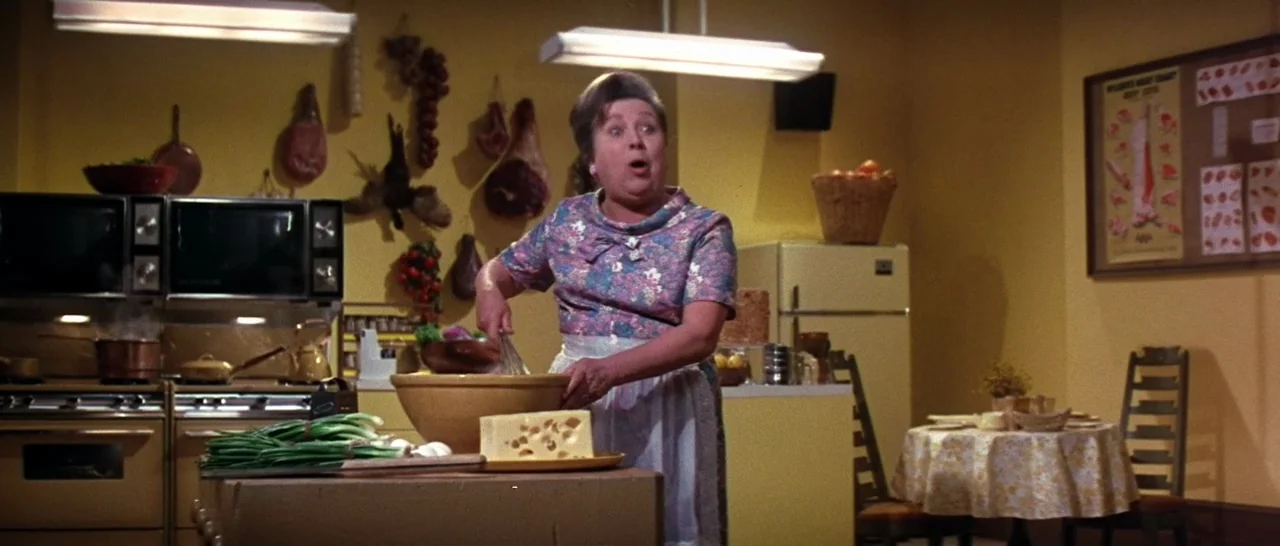 On A Clear Day You Can See Forever (1970) movie still: medium — A retro yellow kitchen features hanging cured meats and a butcher's chart on…