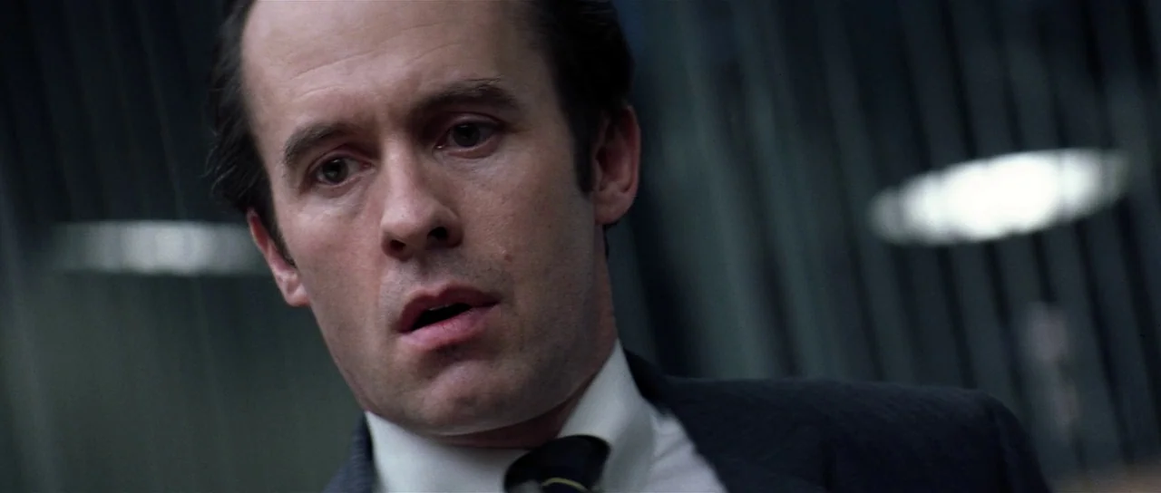 Spy Game (2001) movie still: closeup — A dimly lit professional environment with vertical metallic blinds visible in the background. A man wearing a…