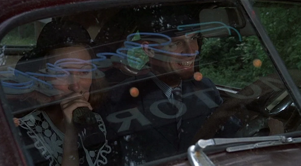 Thieves Like Us (1974) movie still: medium — A man and woman sit inside the front seats of a moving car. Reflections of a blue neon sign are visible…