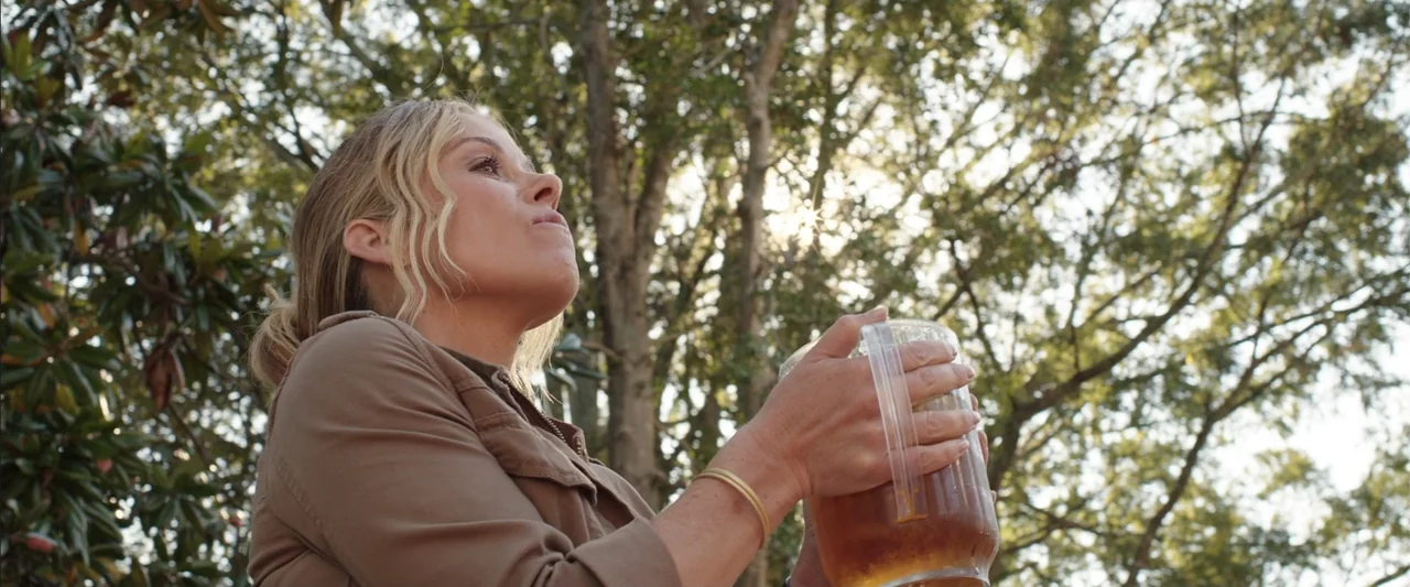 Vacation (2015) movie still: medium — An outdoor area with mature trees and dappled sunlight filtering through the canopy. A woman holds a large…