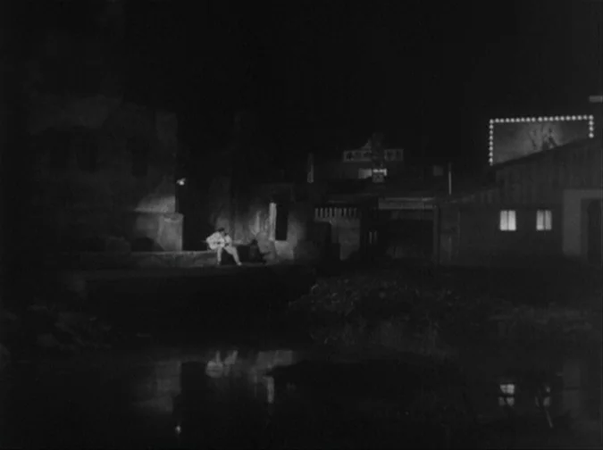 Drunken Angel (1948) movie still: wide — A dark, industrial waterfront at night with a small illuminated structure in distance. A lone man sits on…