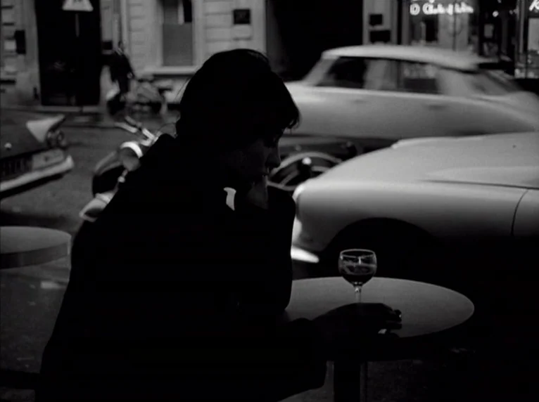 Band of Outsiders (Bande à part) (1964) movie still: medium — A dark night scene at a sidewalk cafe with a vintage car parked nearby. A woman sits…