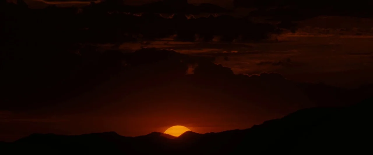Tombstone (1993) movie still: establishing — A vast silhouette of jagged mountain peaks sits against a dark, cloudy twilight sky. The sun is…