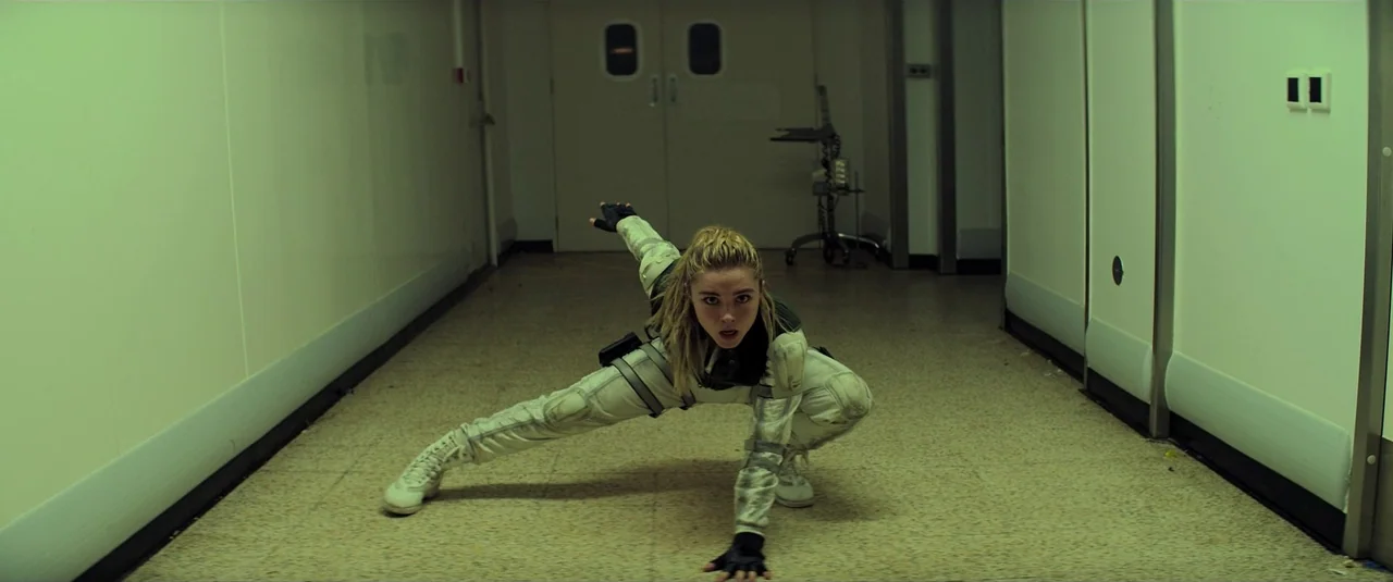 Black Widow (2021) movie still: fullbody — A stark medical hallway with off-white walls and speckled linoleum flooring. A woman in a white tactical…