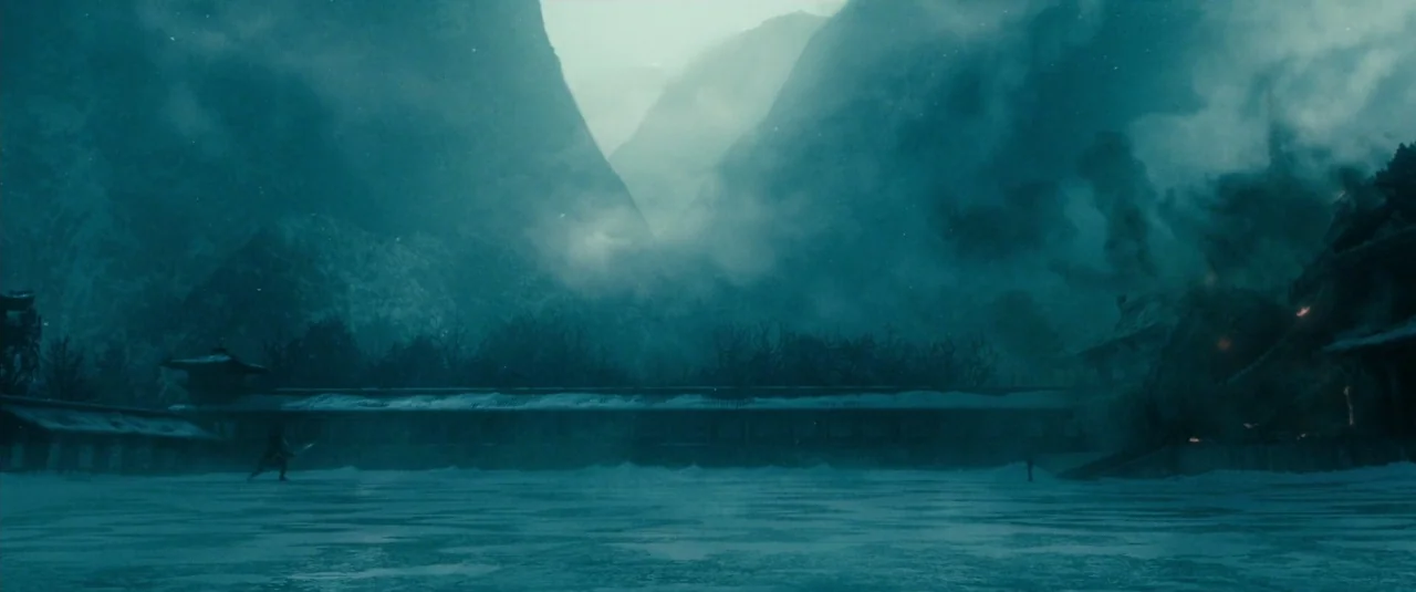 Sucker Punch (2011) movie still: wide — A snowy valley is framed by towering, mist-shrouded dark mountains under an overcast sky. Smoke rises from…