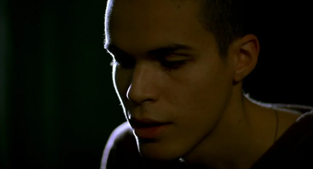 Tiresia (2003) movie still: closeup — A man in his twenties is shown in a dark room with minimal visible detail. The subject keeps his gaze lowered…