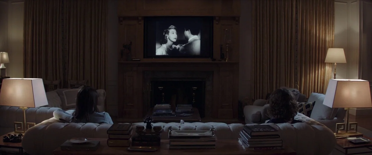 Thoroughbreds (2017) movie still: wide — A dark, wood-paneled living room features a fireplace and a large television screen center. Two individuals…