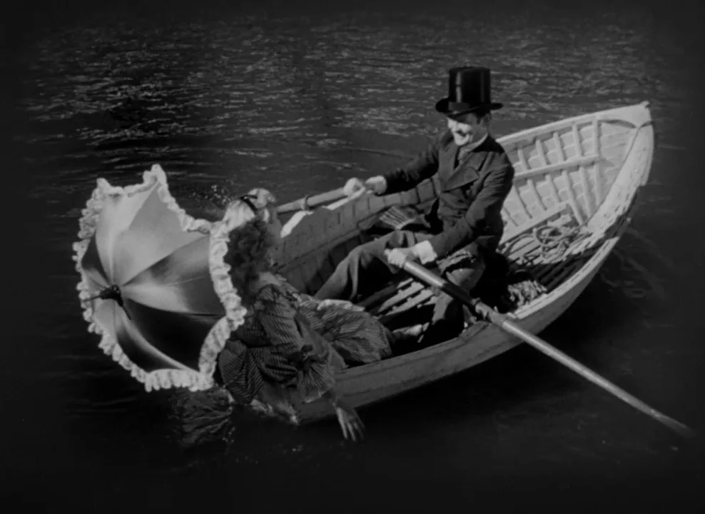The Magnificent Ambersons (1942) movie still: fullbody — Two individuals sit inside a wooden boat floating on a body of dark water. The man operates…