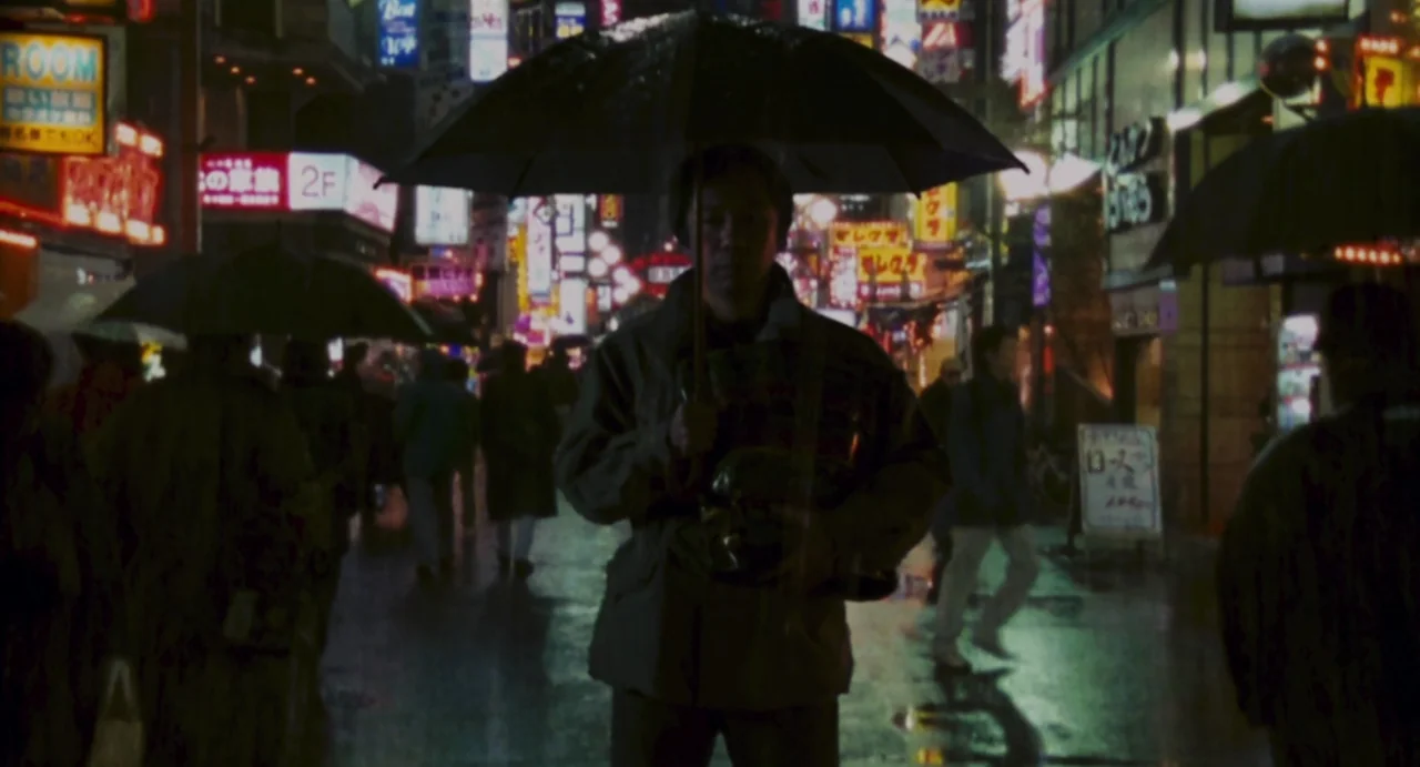 Ichi The Killer (2001) movie still: medium — A rain-slicked city street at night is illuminated by numerous neon signs. A man stands in the center…