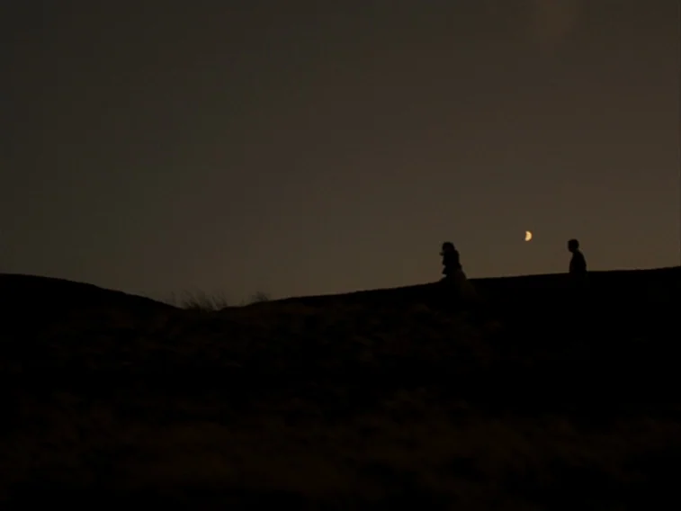 Wuthering Heights movie still: wide — A dark, rolling sandy ridge occupies the foreground under a vast, dark sky. A man and a woman walk along the…